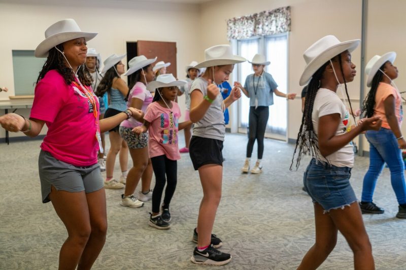 A smiling woman leads a room of young girls doing a country line dance, wearing cowboy hats.