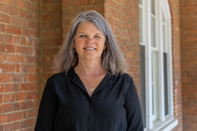 A woman with long gray hair, wearing a black blouse, stands in front of a brick building, smiling for a headshot.