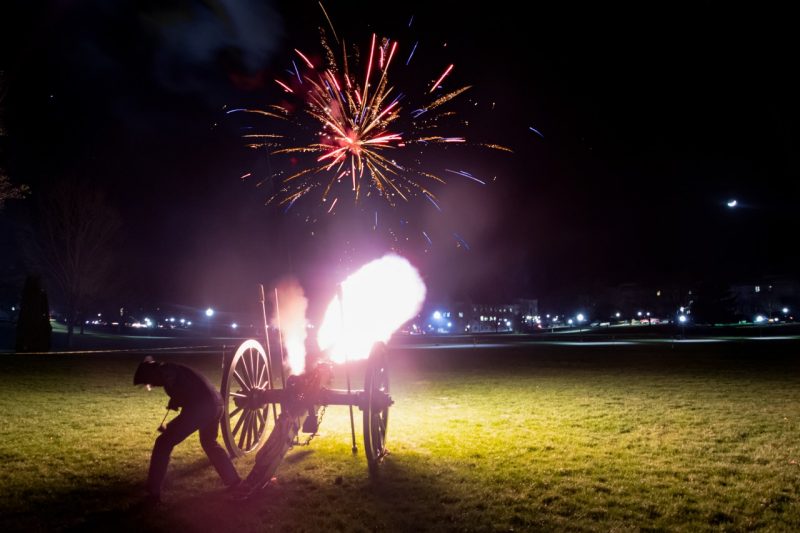 Fireworks in the sky silhouette a cannon being fired by a cadet on a grassy field