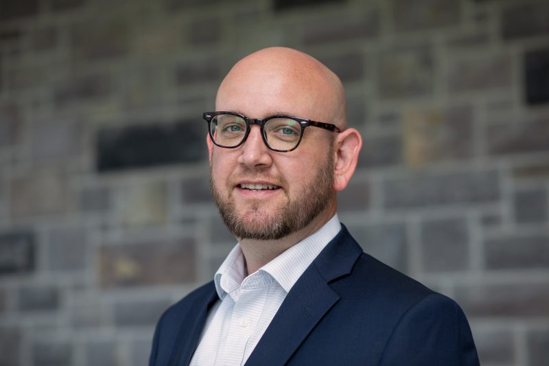 Portrait of Travis Jessee wearing a blue jacket and a white button down shirt smiling.