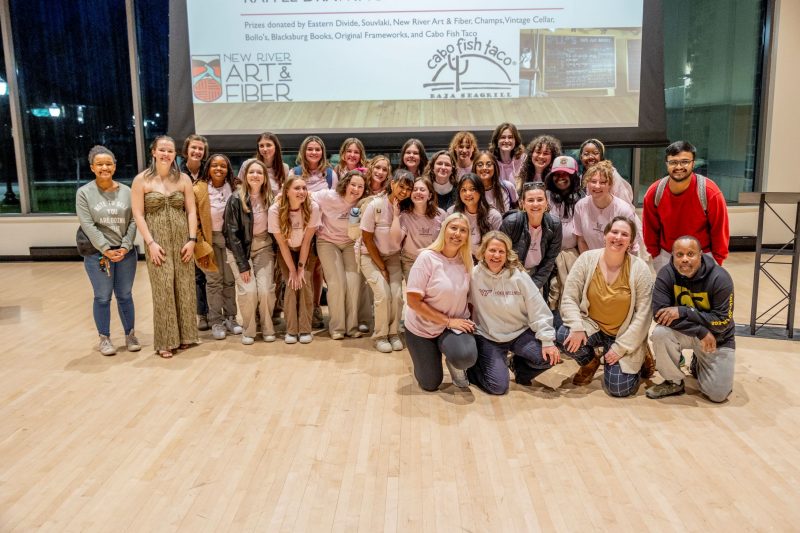 A photo of a group of people posing for a photograph, mostly dressed in khaki pants and light pink t-shirts. Behind them is a screen detailing the businesses that donated prizes to the event as well as some business logos.