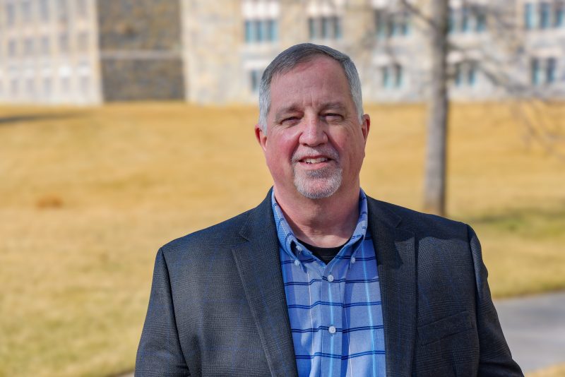 Headshot of Kevin Rice named Clery Compliance Coordinator for Virginia Tech outside with a campus building in the distance.