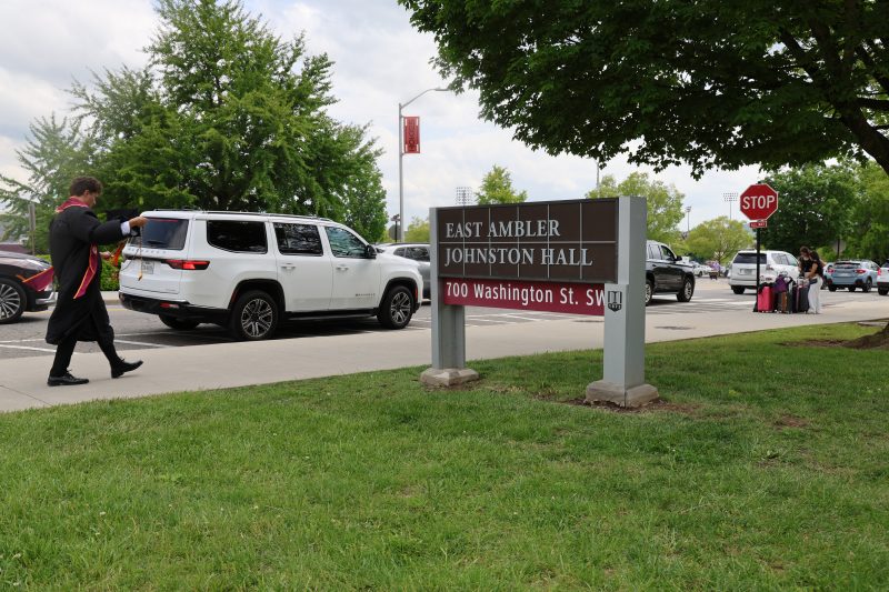 A graduating Hokie walks past the sign for East Ambler Johnston Hall, dressed for a Commencement ceremony. Many cars park along the street for move-out behind him, and two students stand near a stop sign with luggage waiting to be picked up after moving out of their residence hall. 